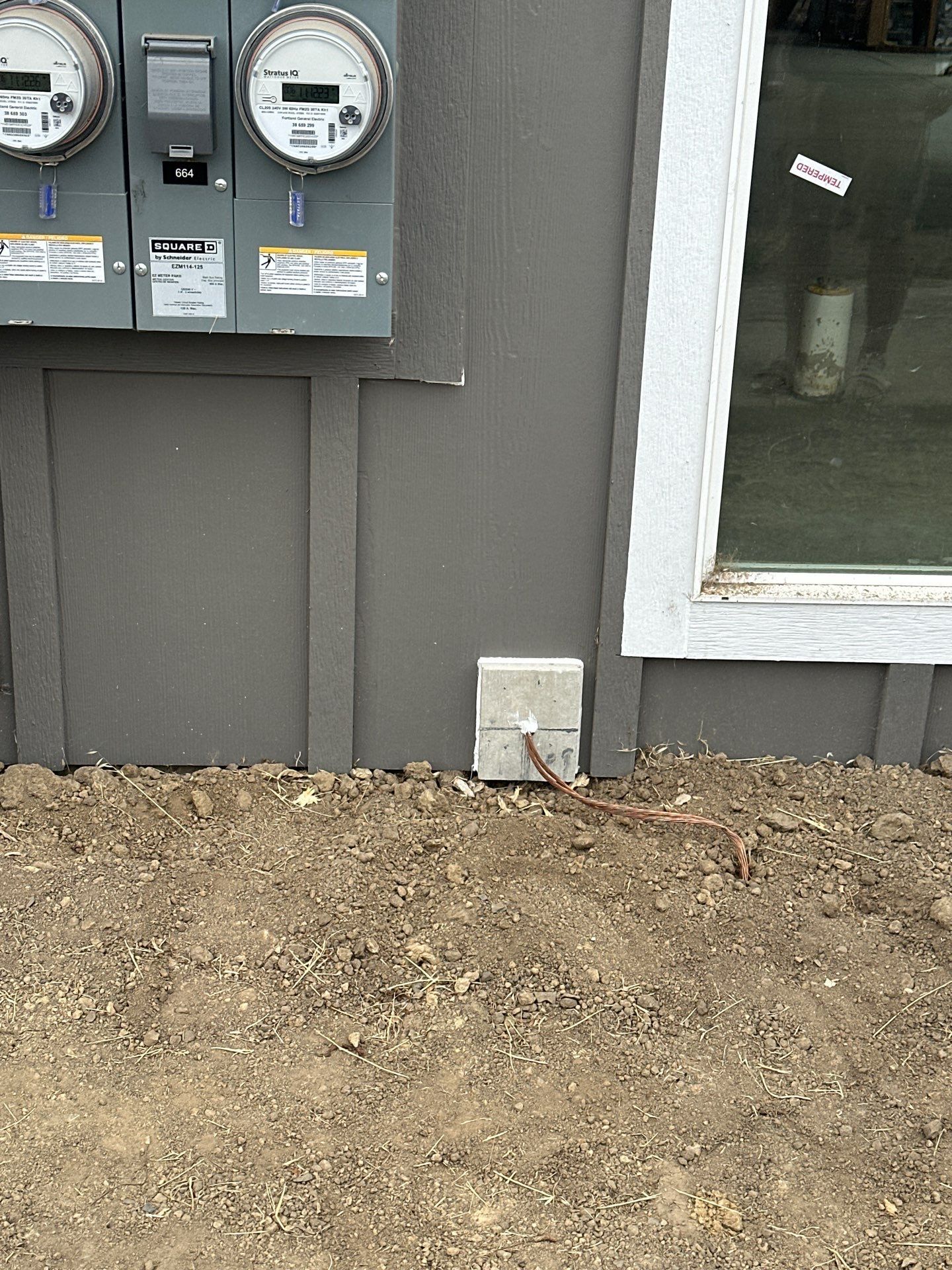 Utility meters on a gray house wall beside a white-framed window and bare dirt ground