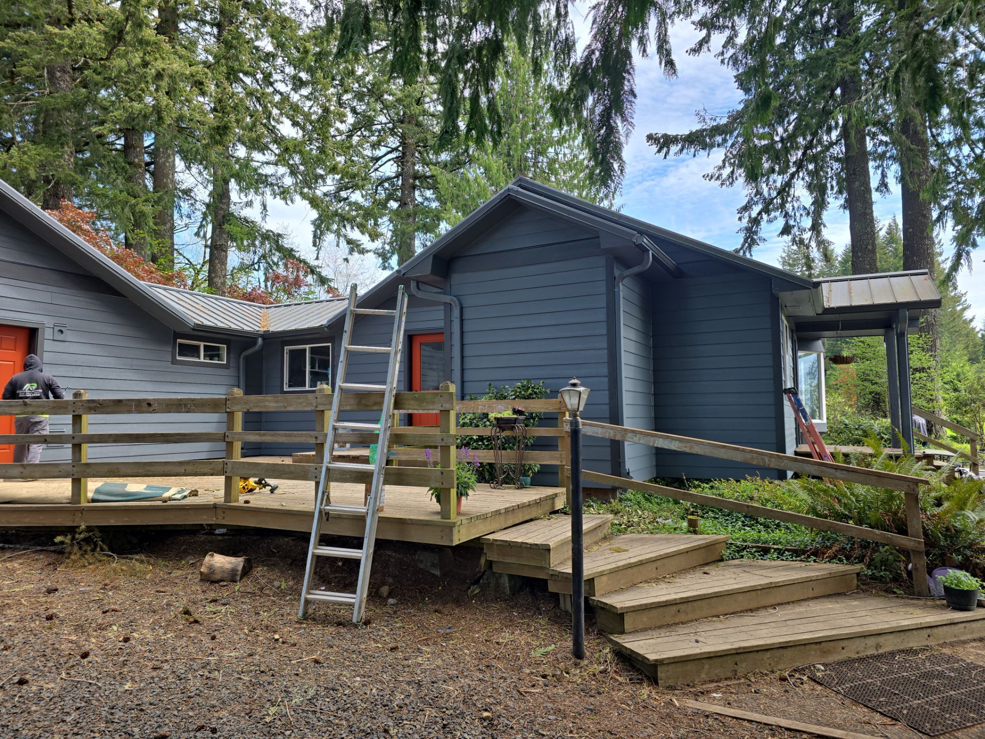 Blue cabin with wooden deck, ladder, and steps in a wooded yard under trees