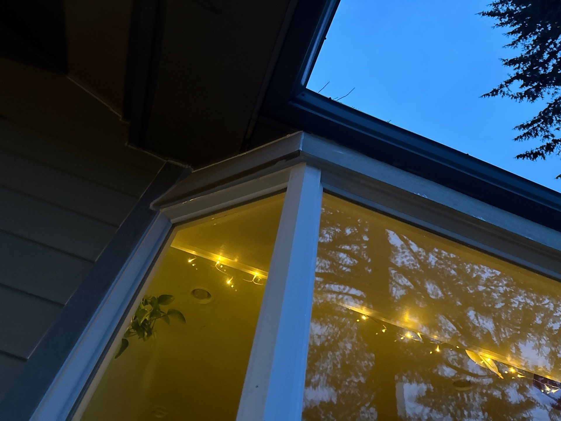 Corner of a house at dusk, with a white bay window glowing yellow under a blue sky.