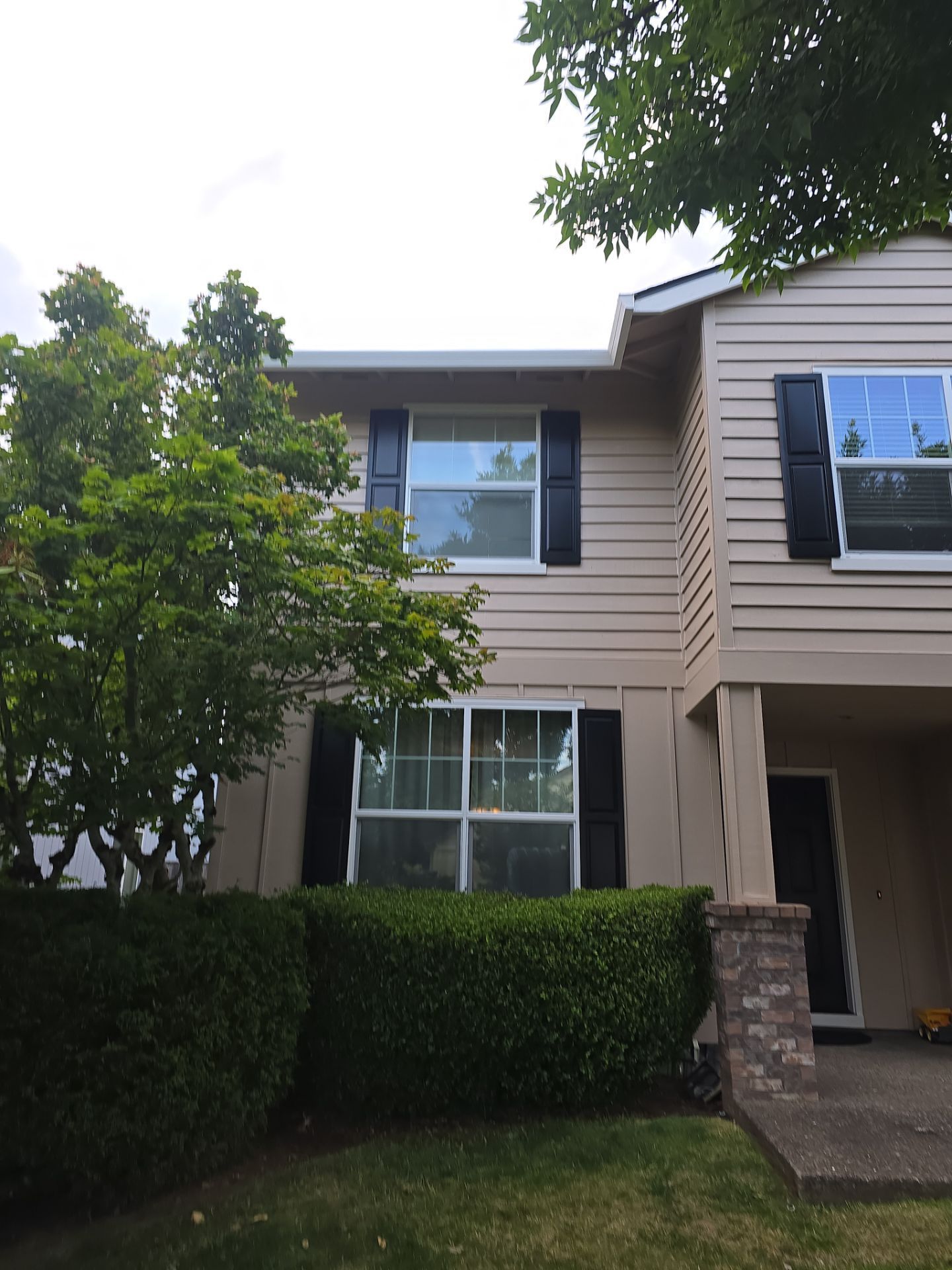 Two-story townhouse with beige siding, dark windows, and a front hedge under a tree canopy