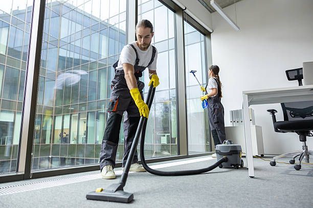 Two people cleaning an office: one vacuuming a carpet, the other dusting. Large windows overlook a city.