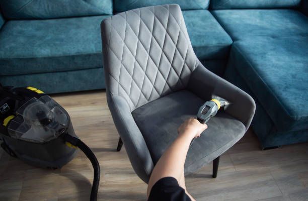 Person vacuuming a gray upholstered chair with a handheld vacuum. Turquoise couch in background.