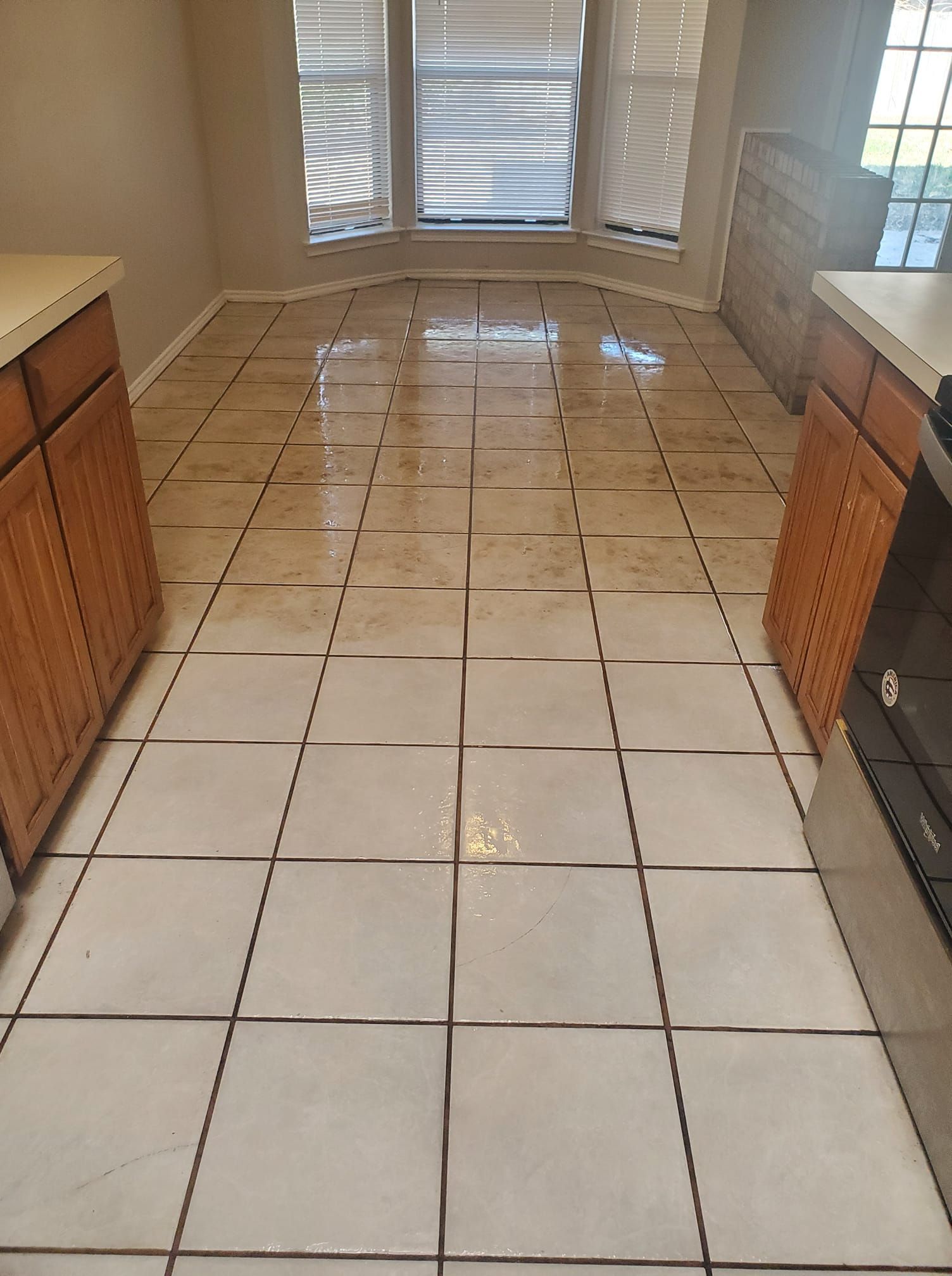 Kitchen floor with light-colored tile and dark grout, cabinets on both sides, and a window in the background.