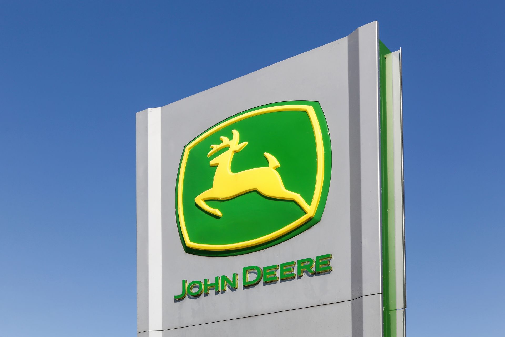 John Deere sign with yellow deer icon, green frame, and green logo text on a white pole.