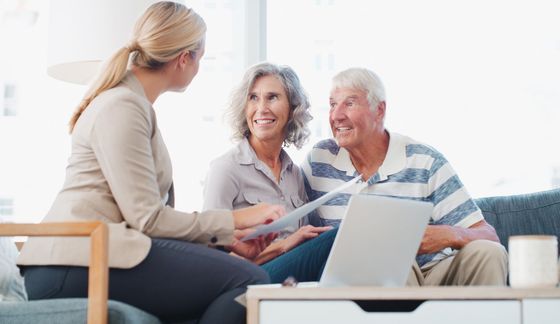An elderly couple is sitting on a couch talking to a woman while looking at a laptop.