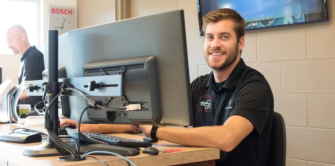 A man smiles while working at a computer in an office. Another person is visible behind him. | Arie Nol Auto Center