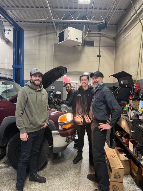 Four people stand in an auto repair shop near a maroon vehicle with its hood open, smiling toward the camera. | Arie Nol Auto Center