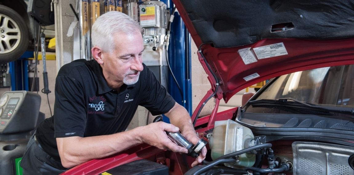 Mechanic working on a car engine in a garage, holding a tool, red car hood open. | Arie Nol Auto Center