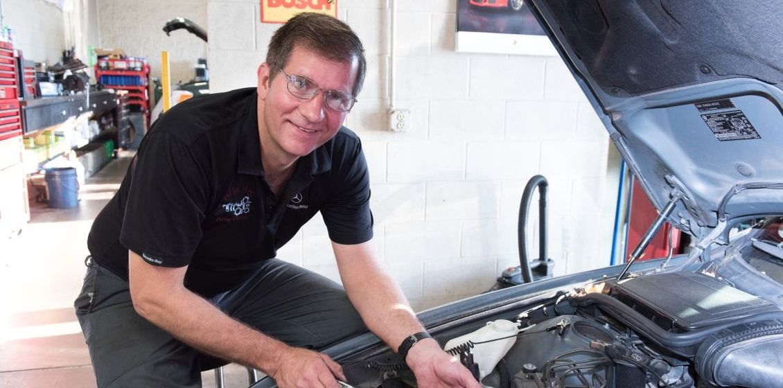Mechanic working on a car engine, smiling. In a shop, wearing black shirt and glasses. | Arie Nol Auto Center