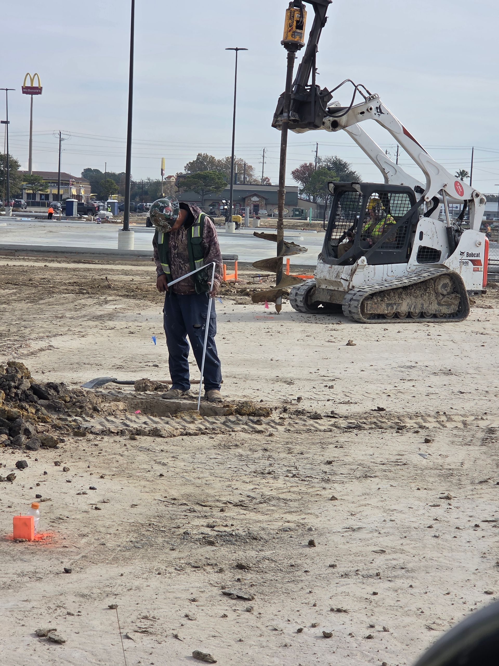 Construction worker observing a drilling operation on a dirt lot, Bobcat equipment in the background.