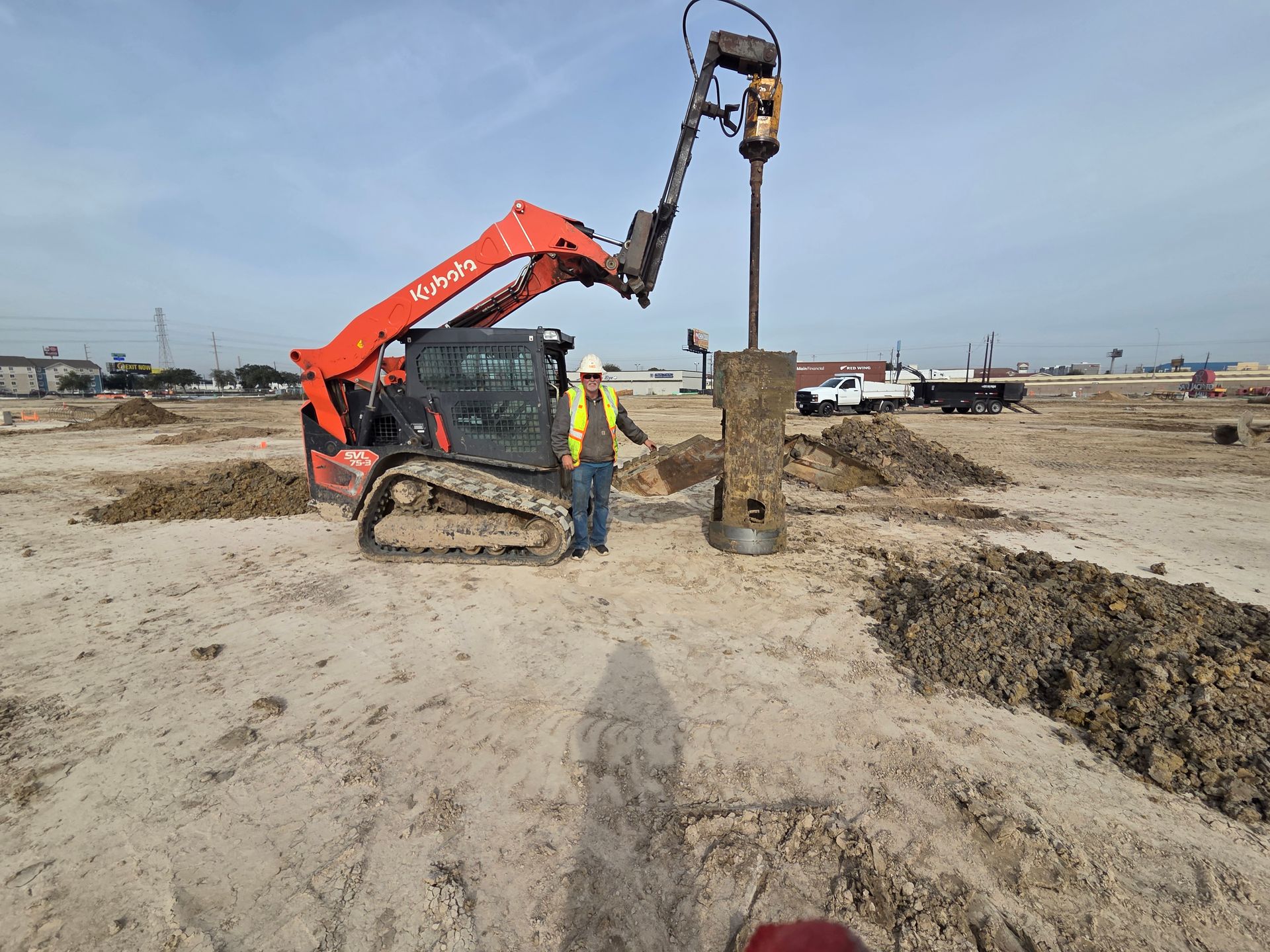 Construction worker beside a Kubota track loader drilling a hole in the dirt.