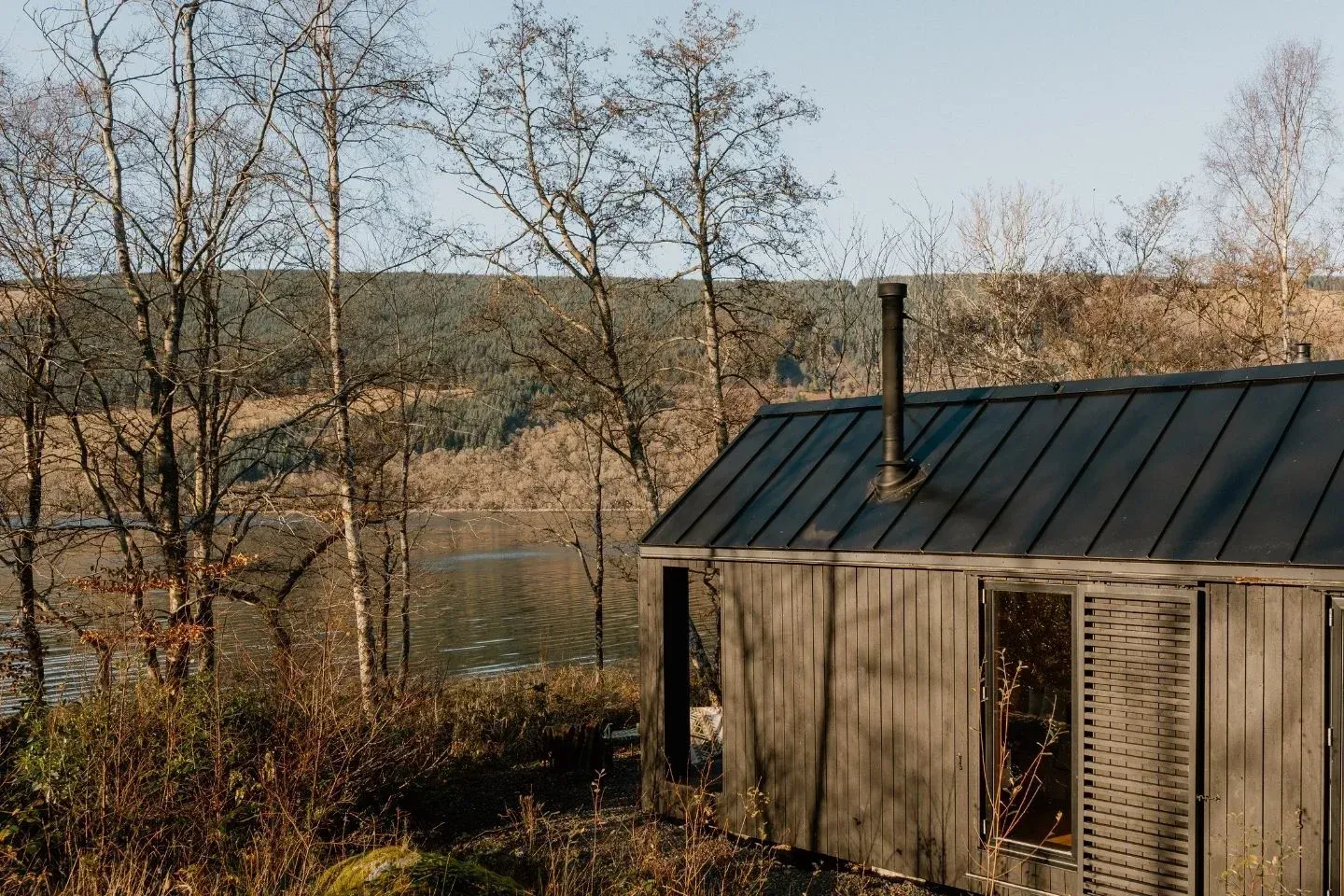 luxury off grid cabin loch fyne, japandi cabin with dark roof and chimney, near a lake with trees.