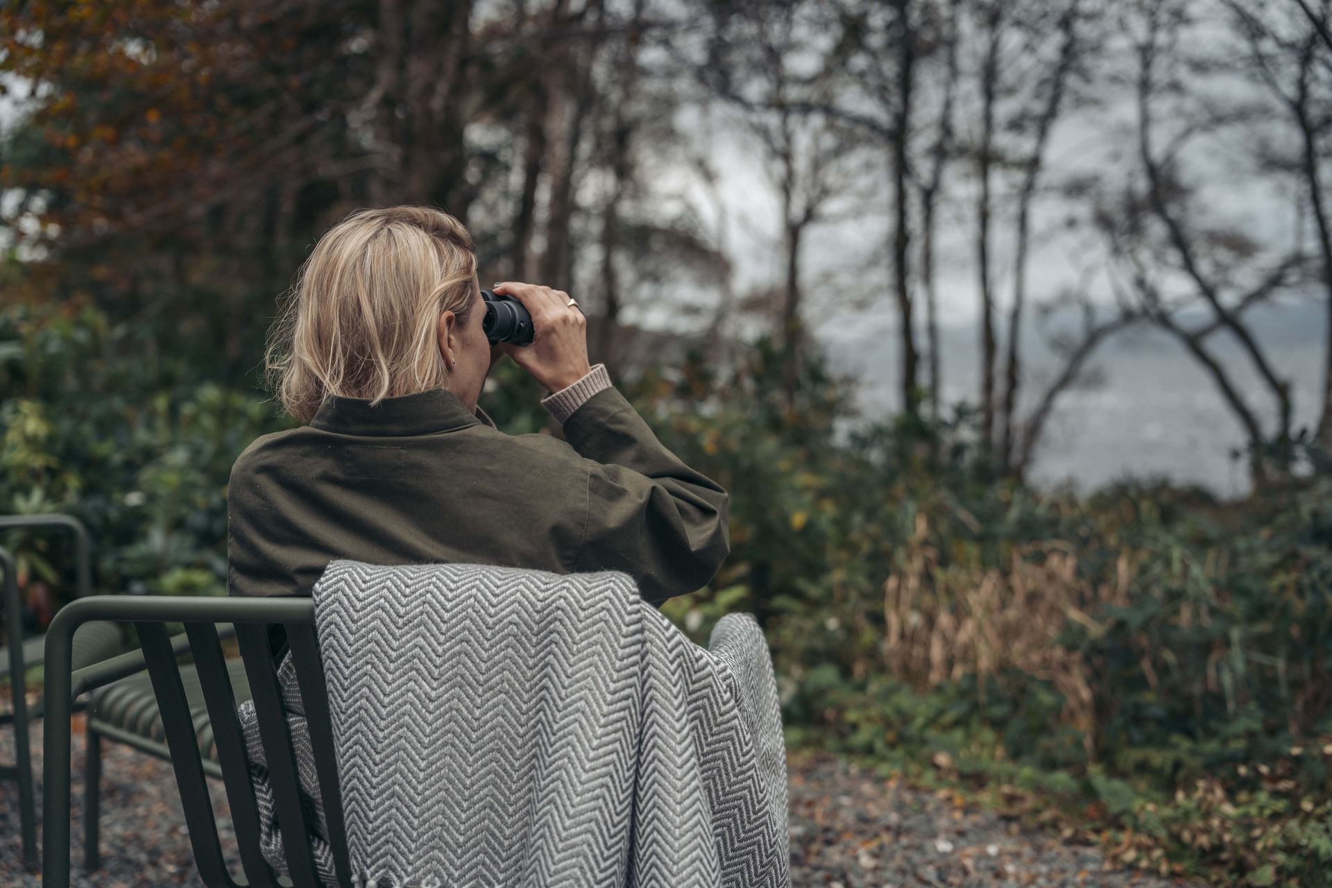 A Kabn guest wrapped in a wool blanket looking through binoculars towards Loch Fyne from the woodland beside the cabin, Argyll, Scotland