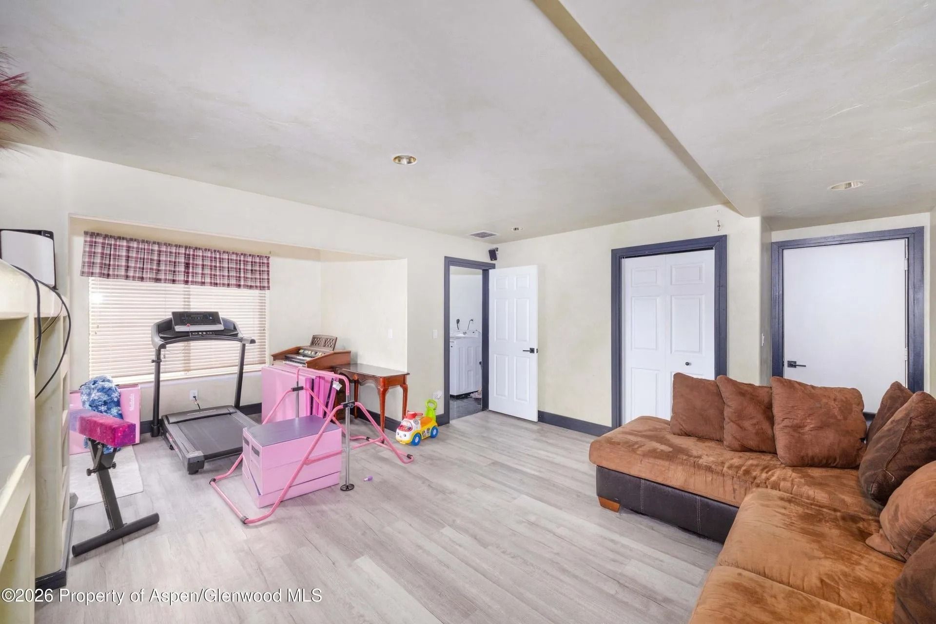 Messy living room with brown sectional, pink baby gear, treadmill, and white walls with blue-trimmed doors
