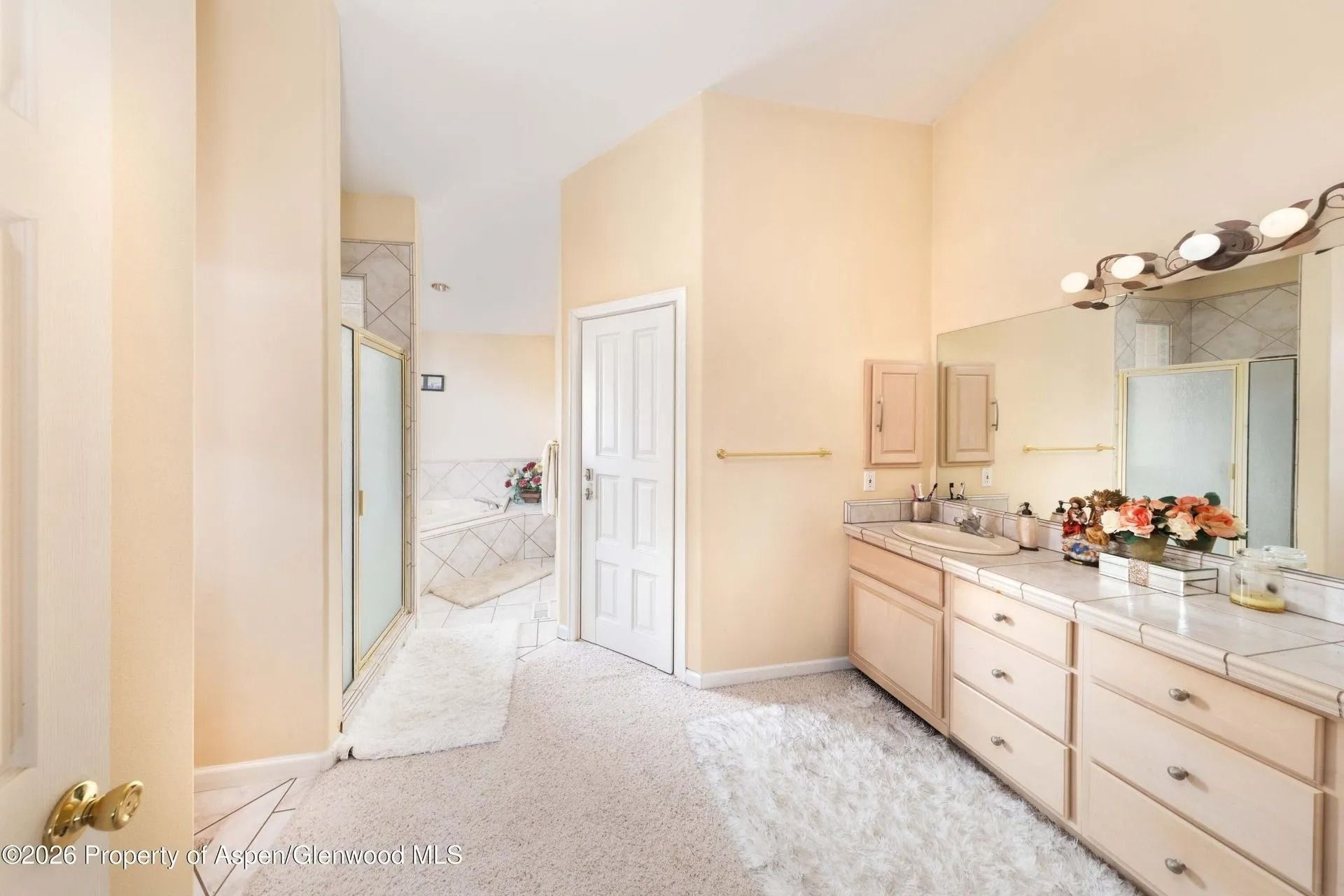 Bright bathroom vanity area with beige cabinets, white walls, and a doorway to a carpeted bedroom.