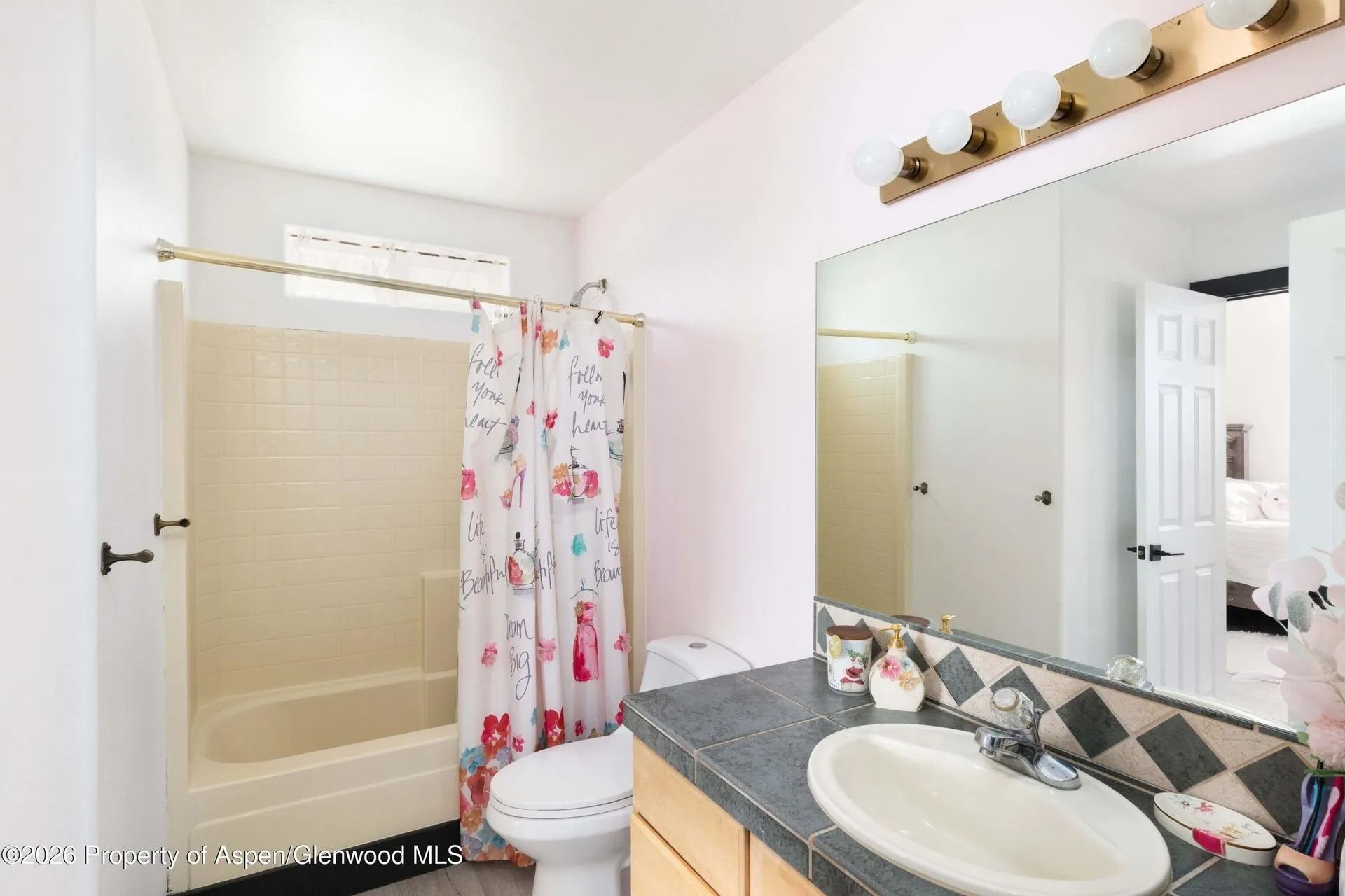 Bright bathroom with beige tub/shower, floral curtain, sink vanity, and large mirror under bulb lights