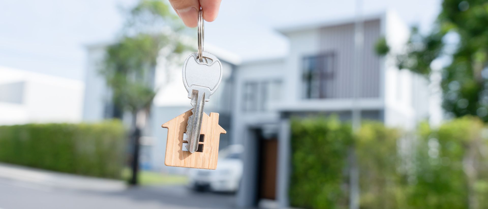 Hand holding a house key in front of a blurred suburban home for sale