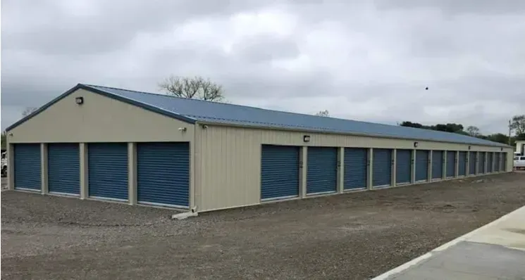 Exterior view of storage units, tan building with blue doors and roof, gravel parking. Exterior view of storage units, tan building with blue doors and roof, gravel parking.