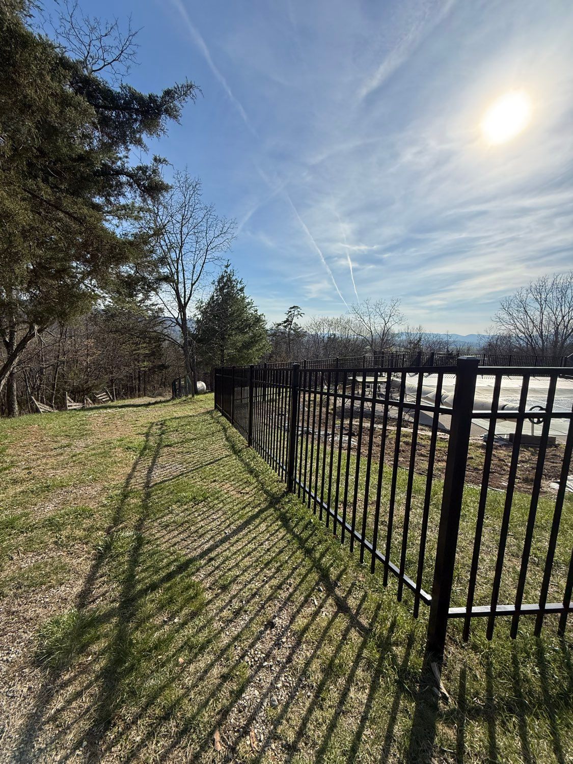 Sunny park path beside black fence casting long shadows over grass and trees