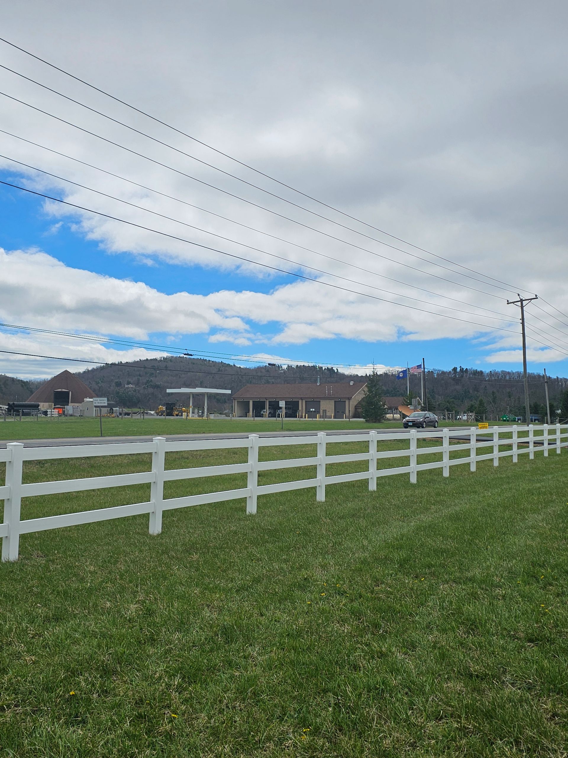 White fence along a green field under a cloudy blue sky with distant buildings and hills