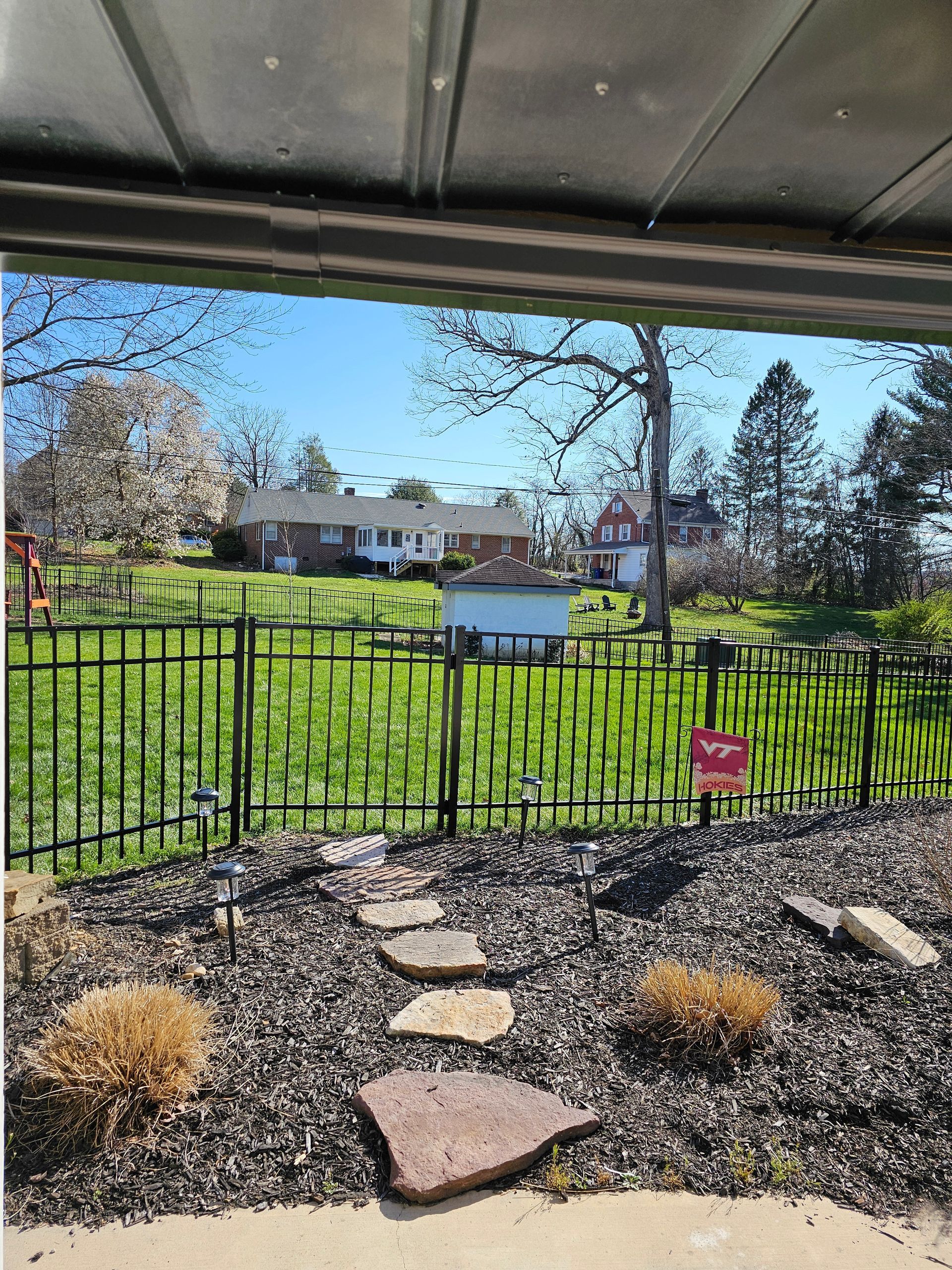 Backyard garden with black metal fence, stepping stones, shrubs, and a house in the distance