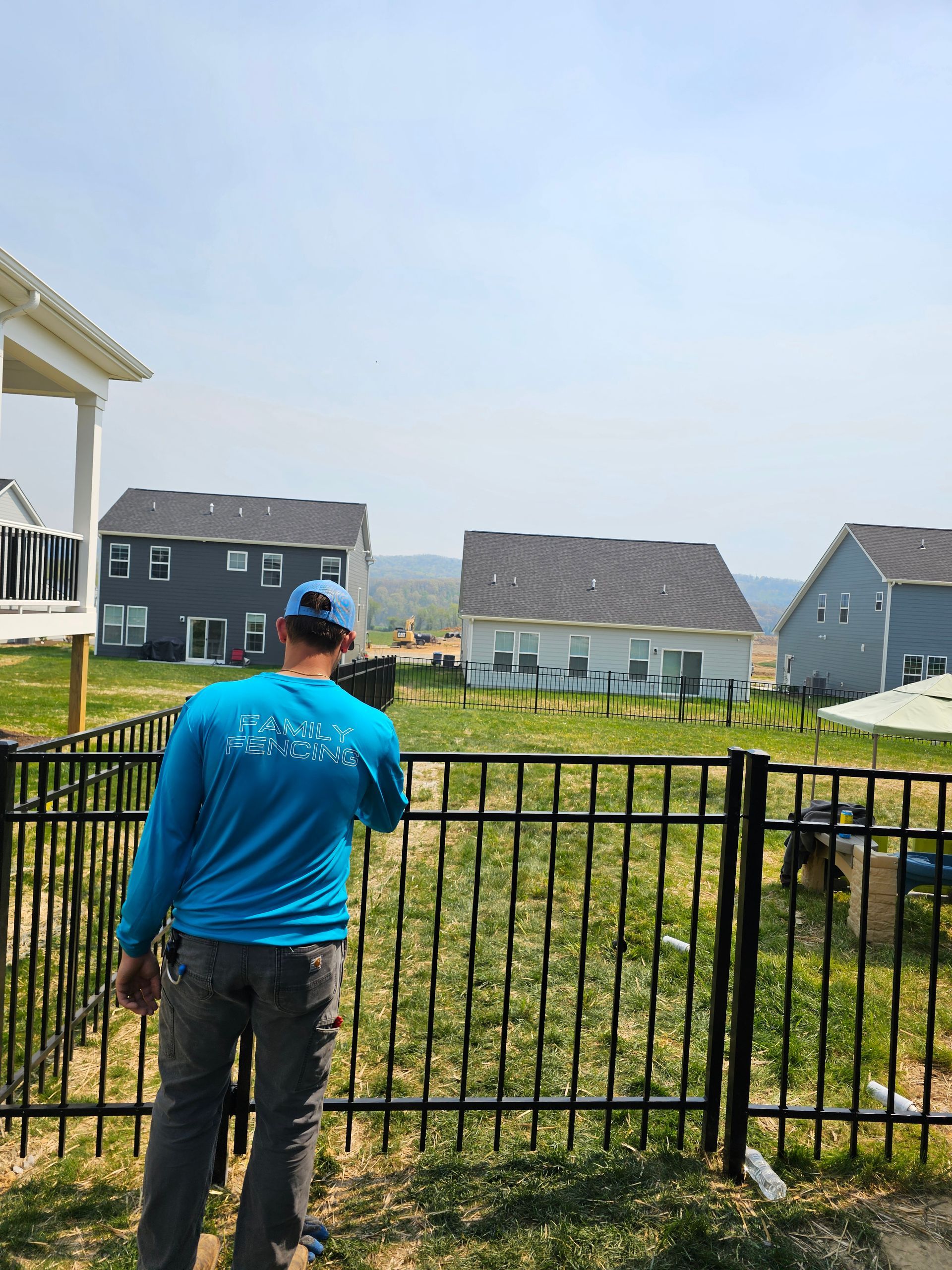 Person in a blue shirt and cap stands by a black fence, looking at houses in a grassy neighborhood.