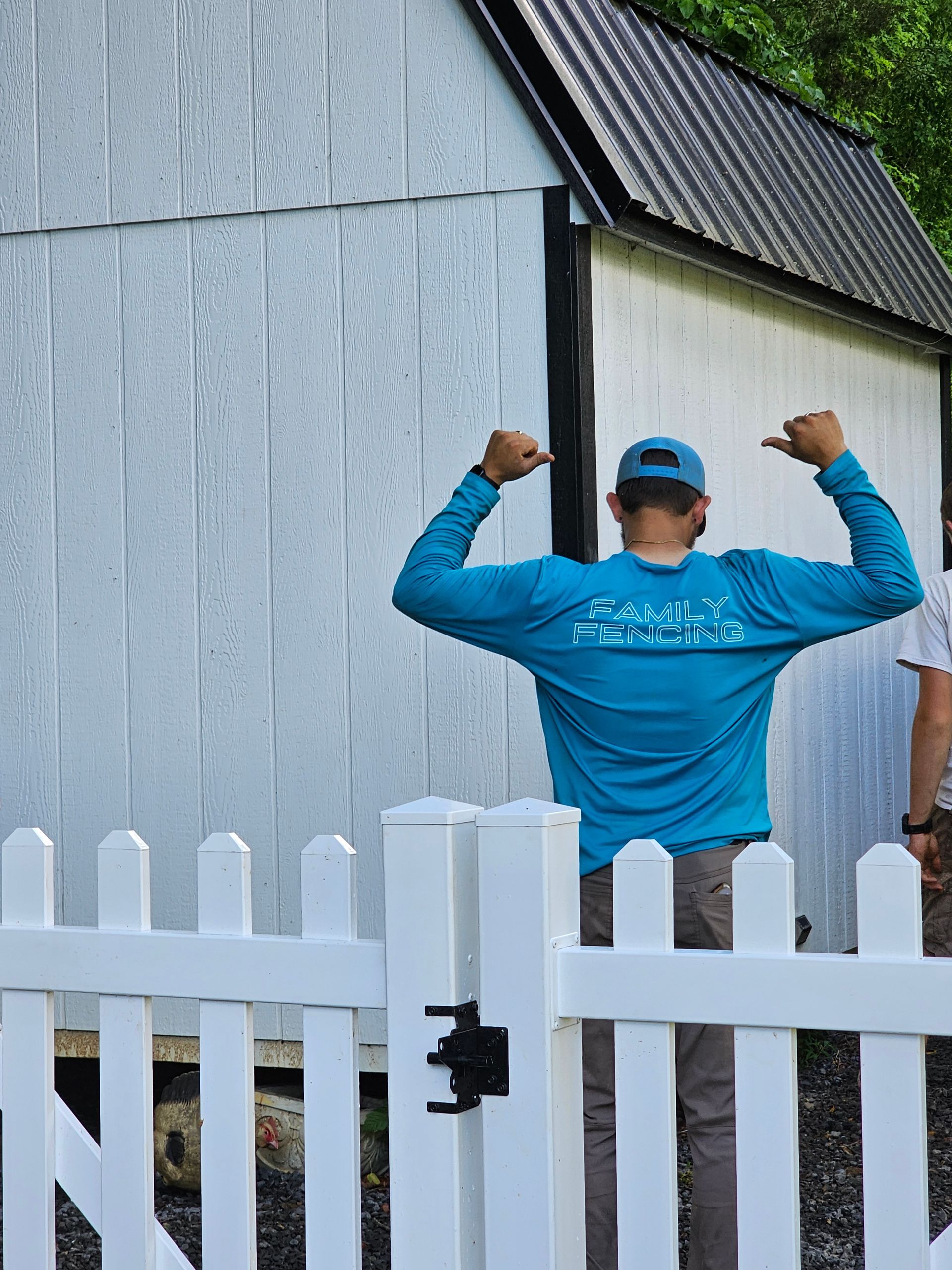 Person in a blue shirt posing with raised arms behind a white fence beside a gray building.