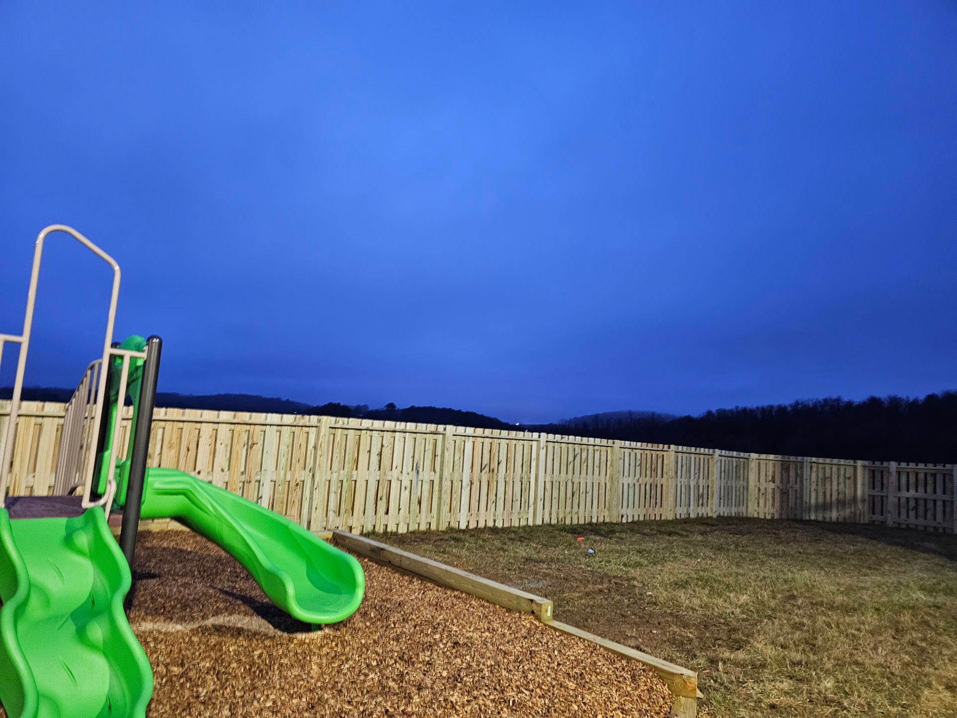 Green playground slide beside a wooden fence under a dark blue evening sky