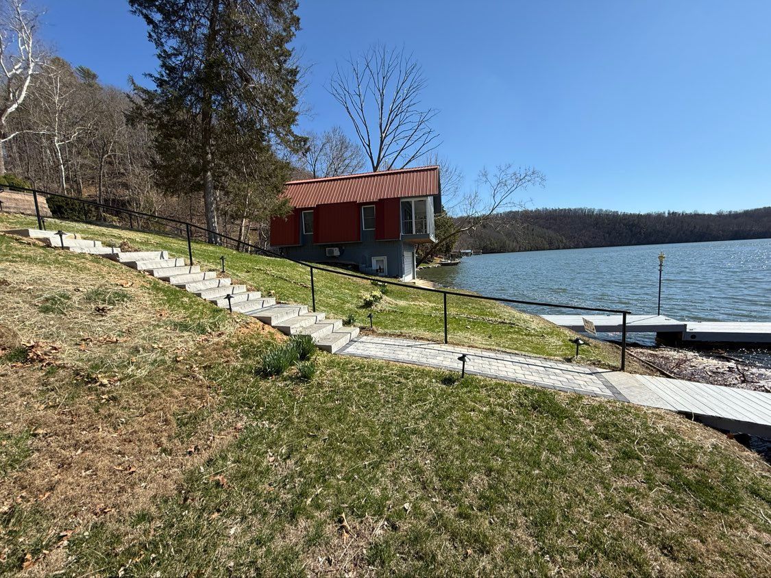 Lakeside path with stone steps, a red-roofed shelter, and calm water under a clear blue sky.