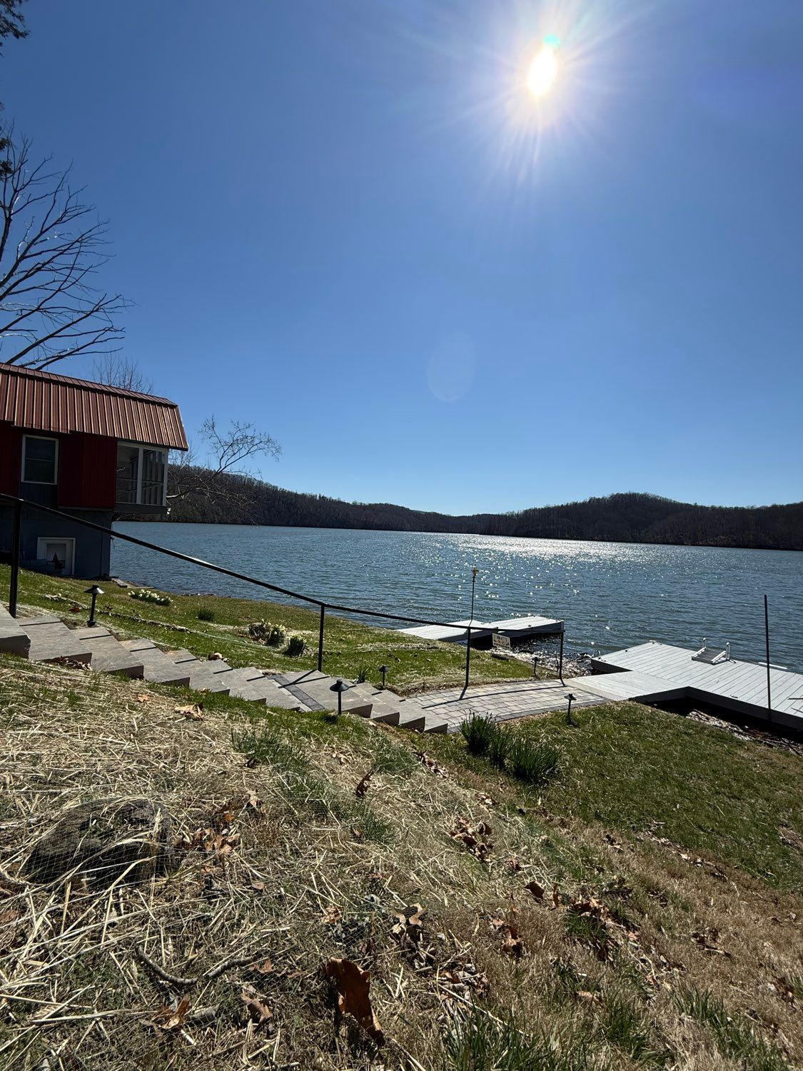 Sunny lakeside dock with a red-roofed cabin, grassy shore, and rippling water under a clear blue sky