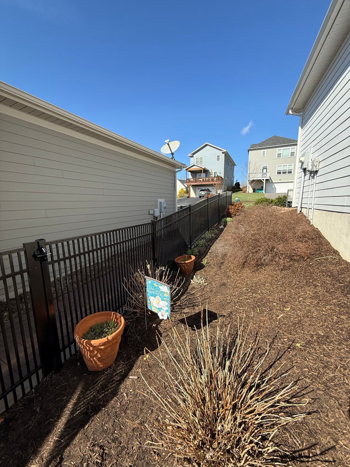 Narrow side yard between beige houses, with a black fence, mulch path, and potted plants under a blue sky.