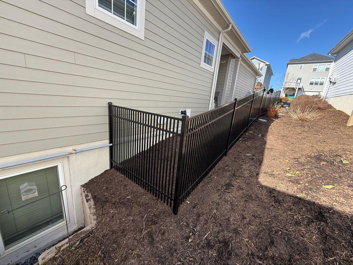 Black metal fence beside beige house along a sloped mulched yard, with neighboring homes in the background