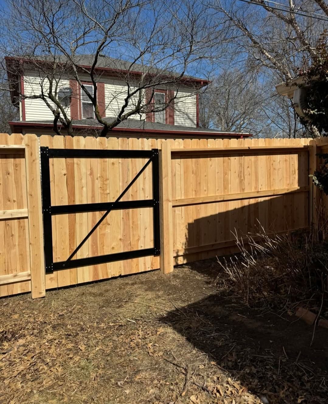 Wooden privacy fence with black gate in a sunny backyard, with a house and bare trees behind it