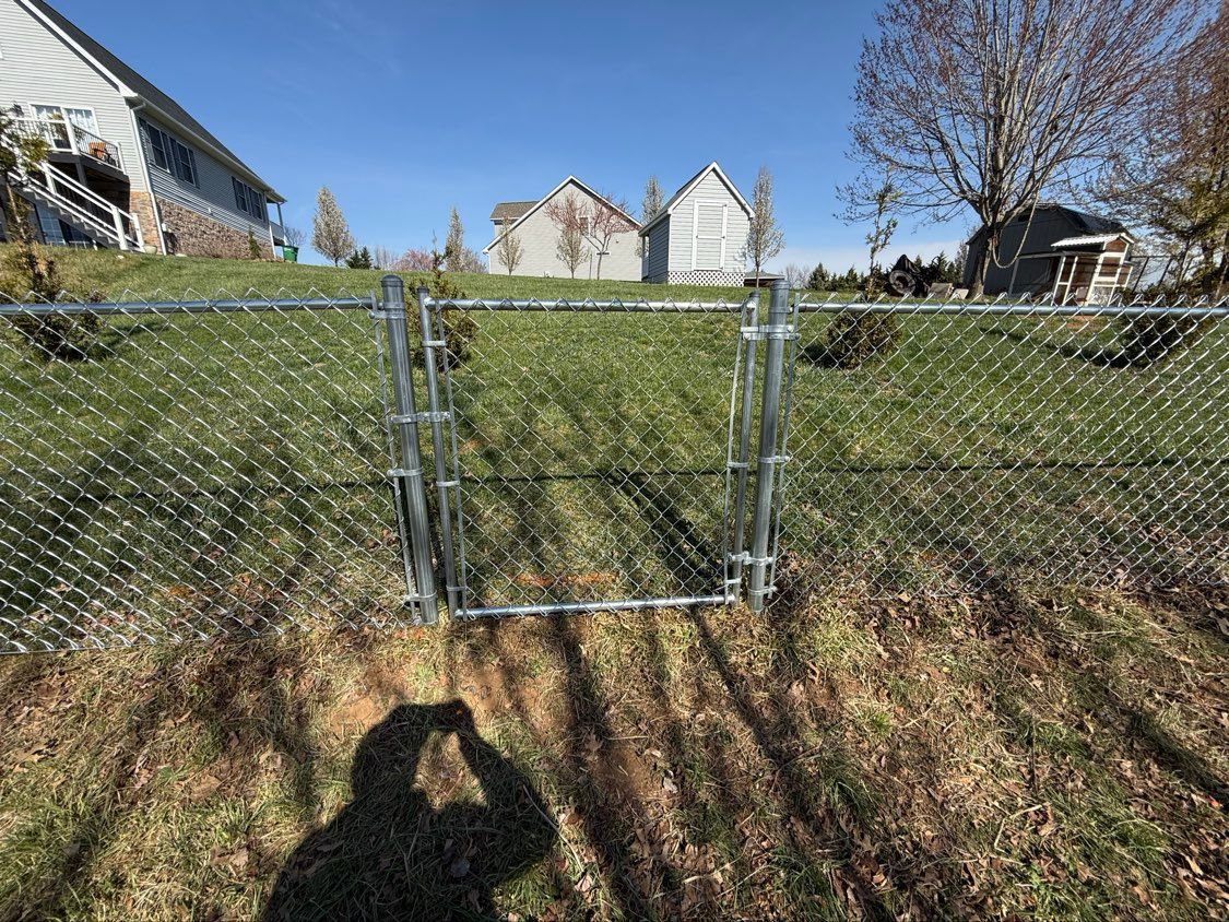 Chain-link fence gate in a grassy yard with houses in the background on a sunny day.