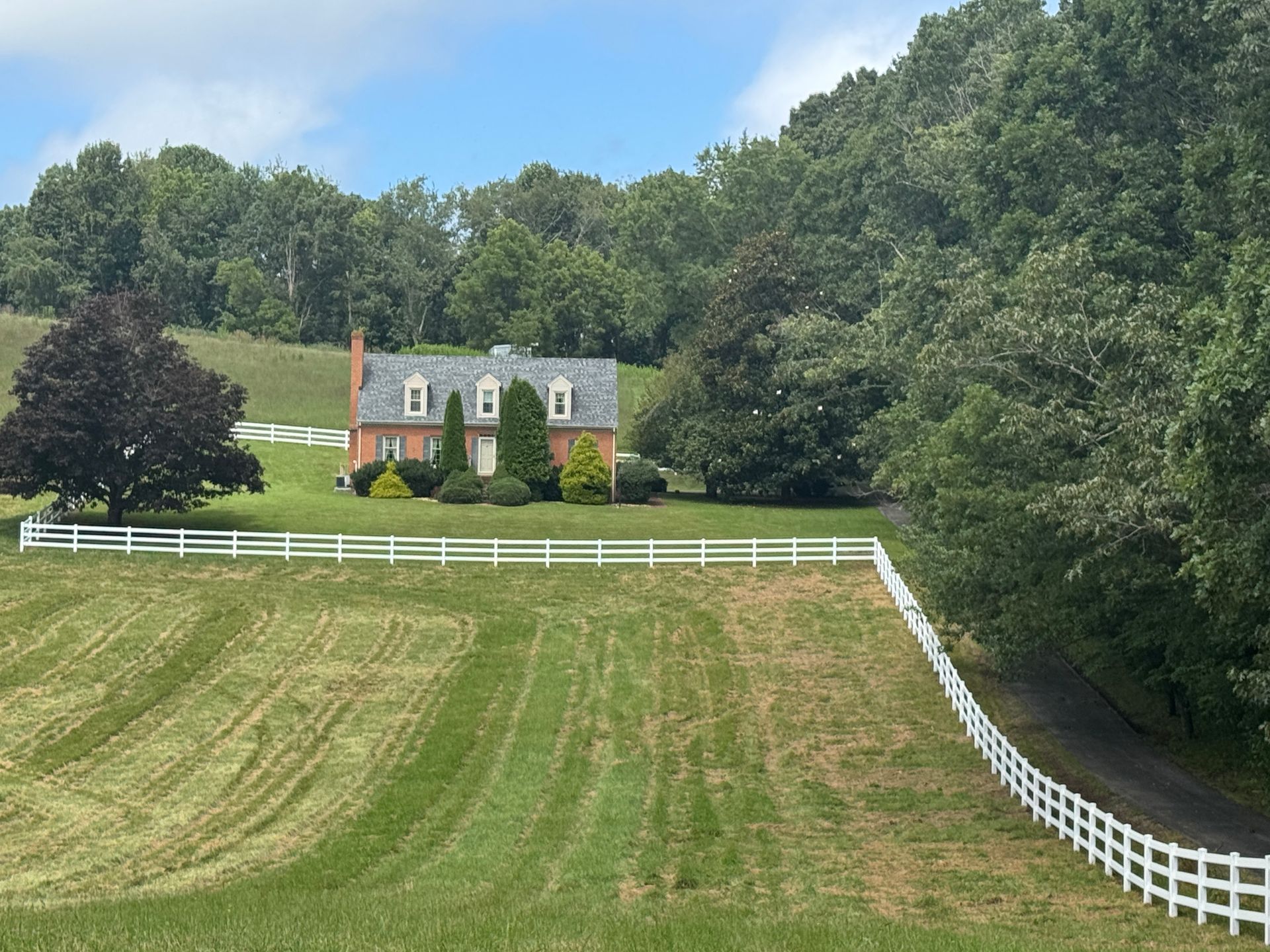 Brick house on a grassy hill with white fence, surrounded by trees under a blue sky