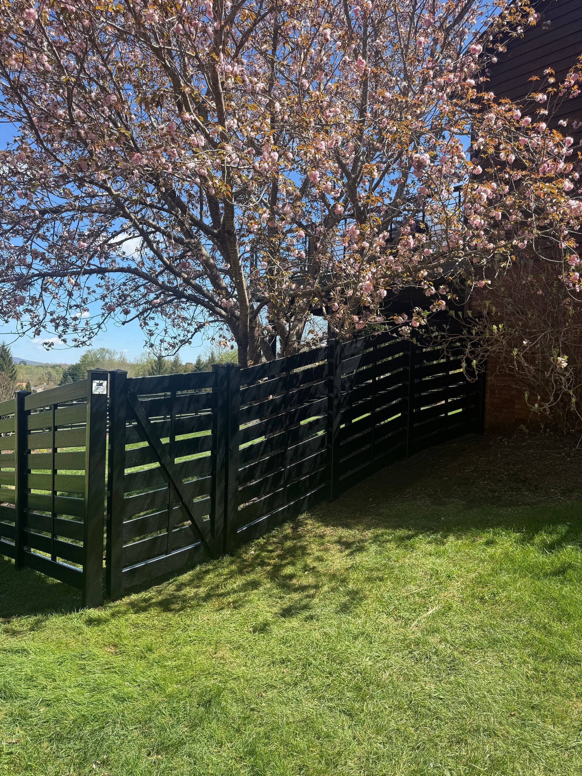 Black wooden fence beside a grassy slope under a blooming tree on a sunny day