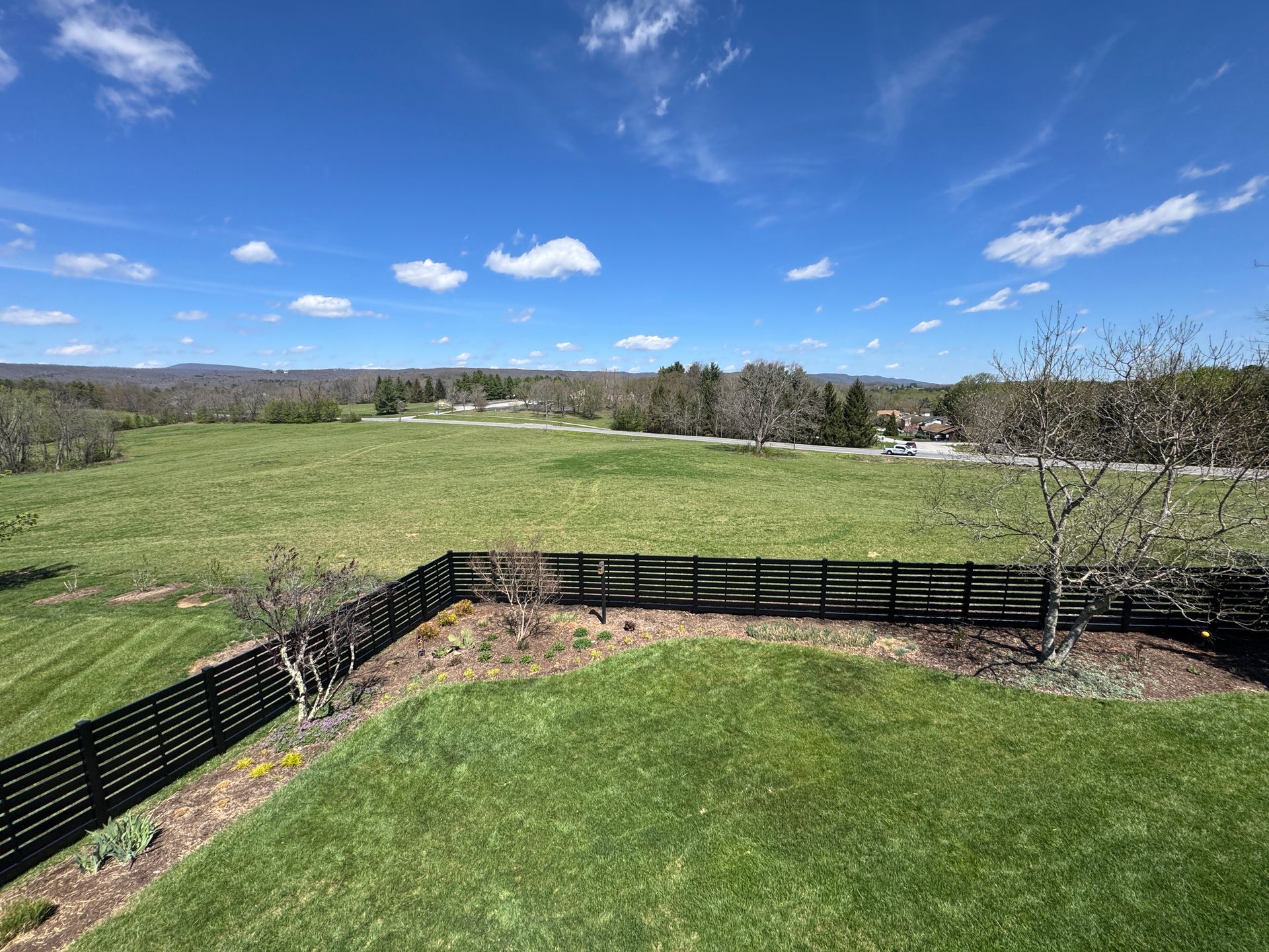 Wide green field with black fence under a bright blue sky with scattered clouds.