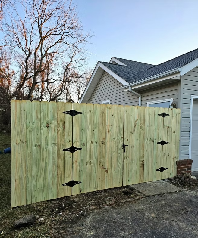 Wooden privacy gate beside a house, with black hinges and bare trees behind it