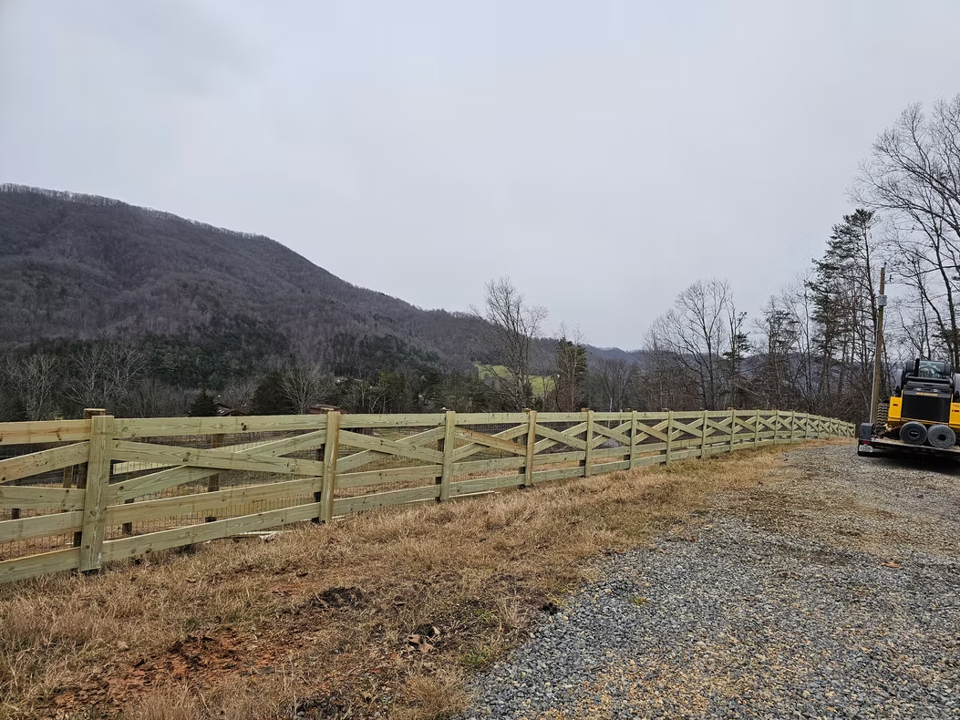Wooden fence along a gravel path beside a wooded hillside, with an excavator at right under a cloudy sky.