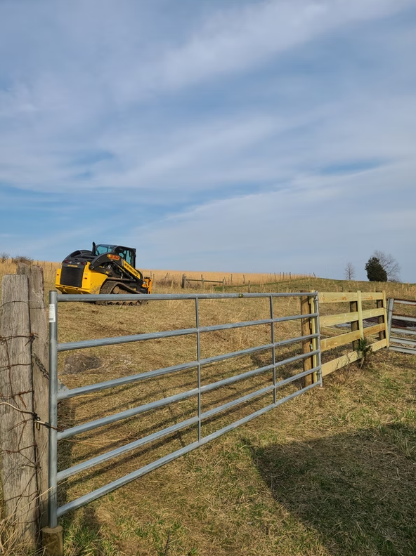 Metal farm gate in a grassy field with a skid-steer loader in the background under a cloudy sky