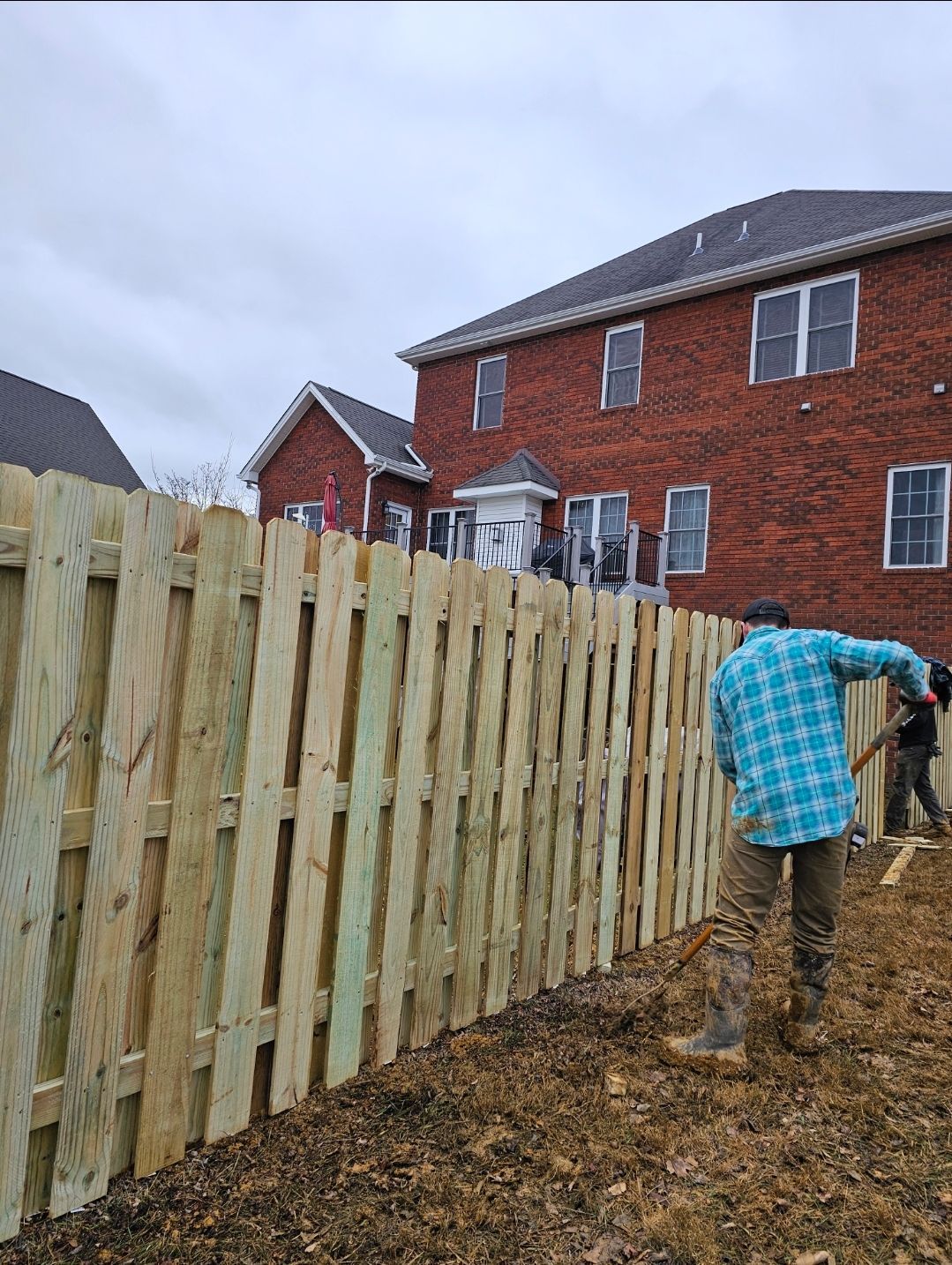 Worker painting a wooden fence beside a red brick house on a cloudy day