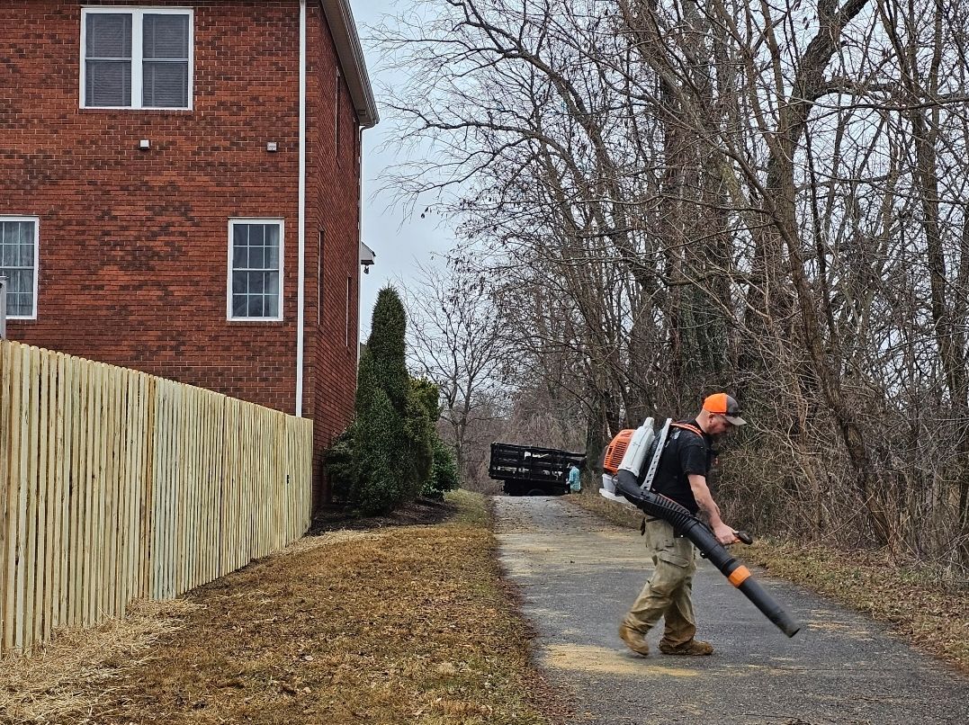 Person using a leaf blower on a wooded path beside a red brick house on a gray day