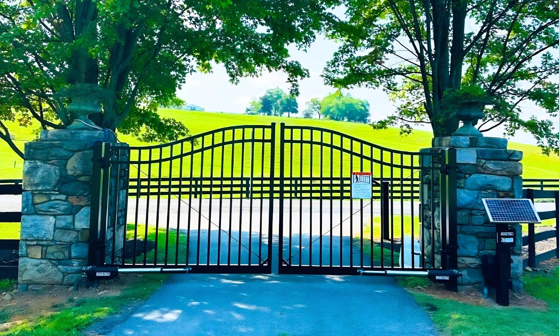 Closed black iron gate between stone pillars at a park entrance, with green trees and lawn behind it.