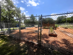 Chain-link gate at a fenced playground with wood chips, trees, and a blue sky