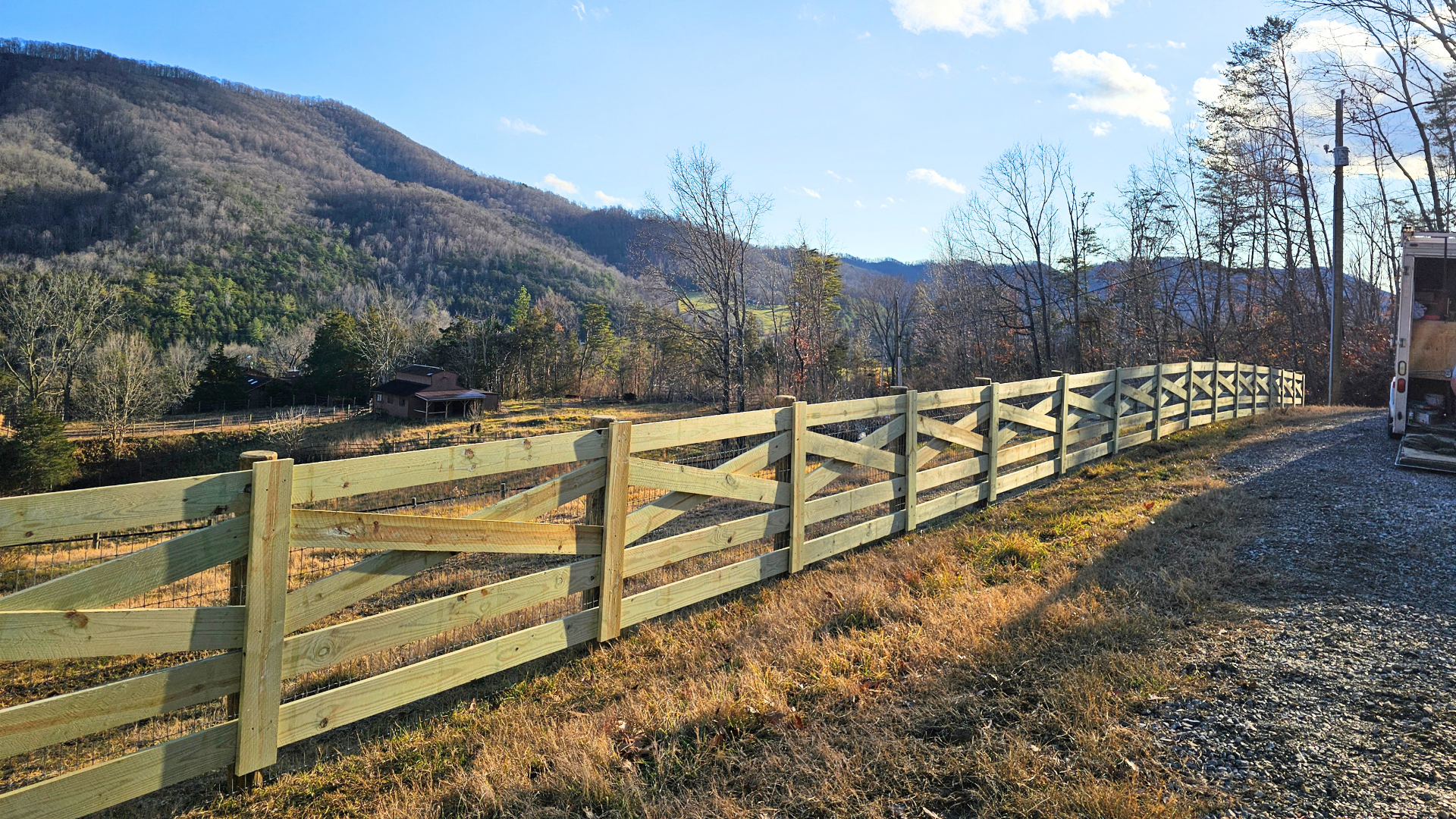 Wooden fence beside a sunny rural road, with mountains and bare trees in the background.