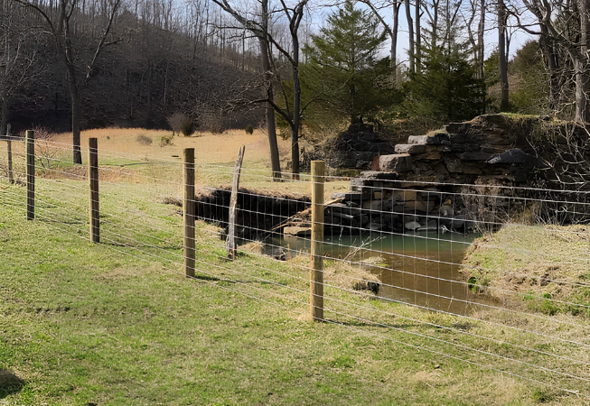 Fenced grassy field with a small pond and rocky waterfall beside trees
