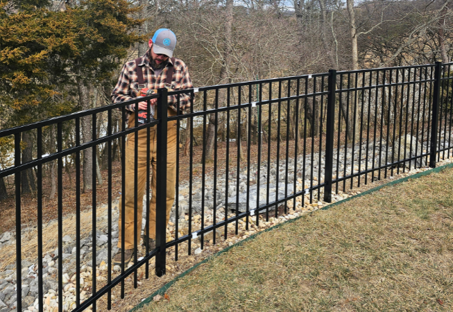Person in a blue cap working beside a black metal fence along a grassy yard.