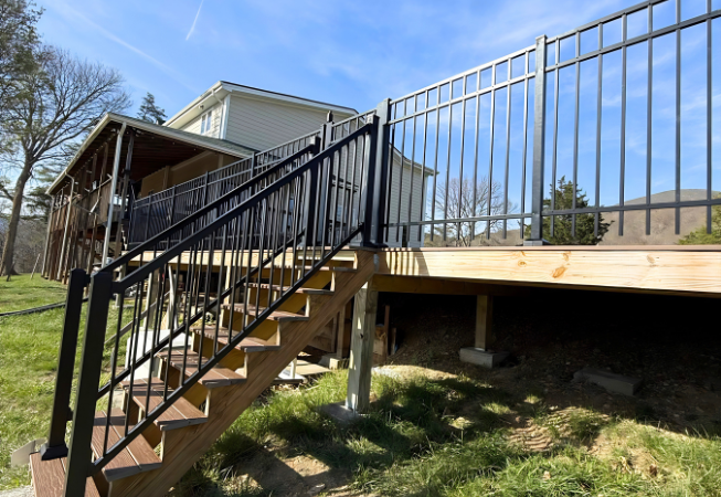 Outdoor stairs with black railings leading to a raised wooden deck beside a house on a sunny day