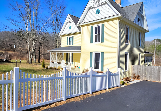 Yellow two-story house with white trim, front porch, and white picket fence beside a driveway