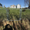 Chain-link fence with open gate overlooking grassy slope and houses under a blue sky
